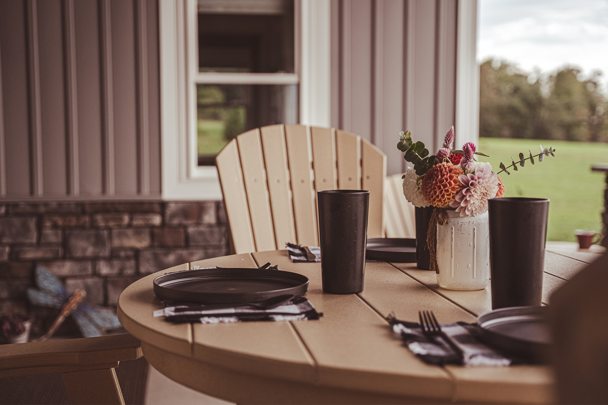 An outdoor beige table with black plates and cups is set on a porch, featuring a mason jar with vibrant flowers as a centerpiece. The setup is in front of a beige and stone wall, with a green landscape visible through a nearby window.