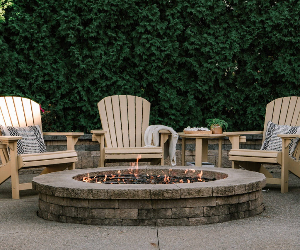 A cozy outdoor seating area with four Adirondack chairs arranged around a stone fire pit, accented with decorative pillows and a side table, creating a warm and inviting backyard retreat with stylish outdoor furniture.